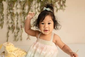 Little girl waving her hand full of cake and smiling as she eats cake for her cake smash photo session. She's in a rainbow dress and smiling at the Philadelphia photographer.